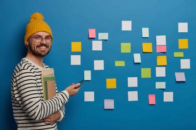 Sideways shot of happy bearded hipster student wears yellow hat and striped jumper, holds notepads and papers, uses mobile phone, stands against blue wall with colored adhesive reminder notes